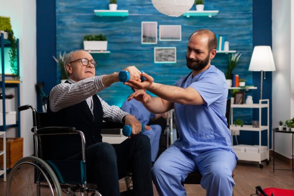 Practitioner man doctor helping retiree senior male in wheelchair to do physiotherapy strength exercise Practitioner man doctor helping retiree senior male in wheelchair to do physiotherapy strength exercise using dumbbell. Social nursing services at home. Healthcare recovery. Support worker helper